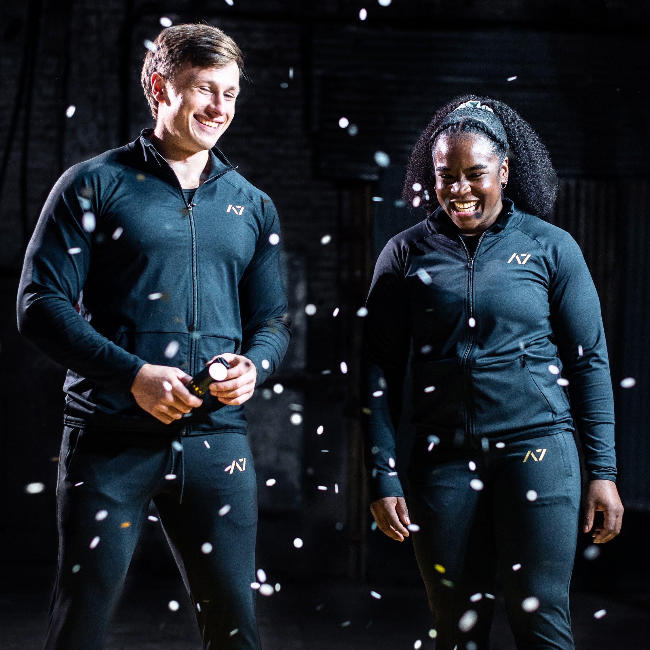 Male and female athletes wearing the Defy Jacket – Gold Standard, standing side by side to showcase the unisex fit, gold silicone logo detailing, and tapered design.